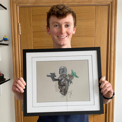Tom Mothersole holding framed realistic coloured pencil drawing of LEGO Mandolorian and Grogu Minifigures
