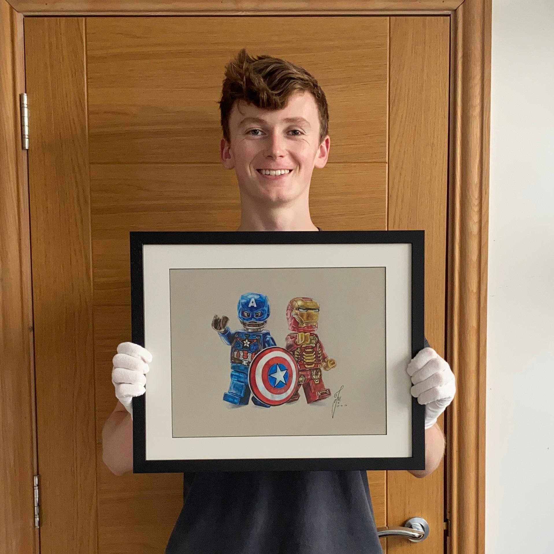 tom mothersole holding framed realistic coloured pencil drawing of the LEGO Captain America and Iron Man Minifigures 