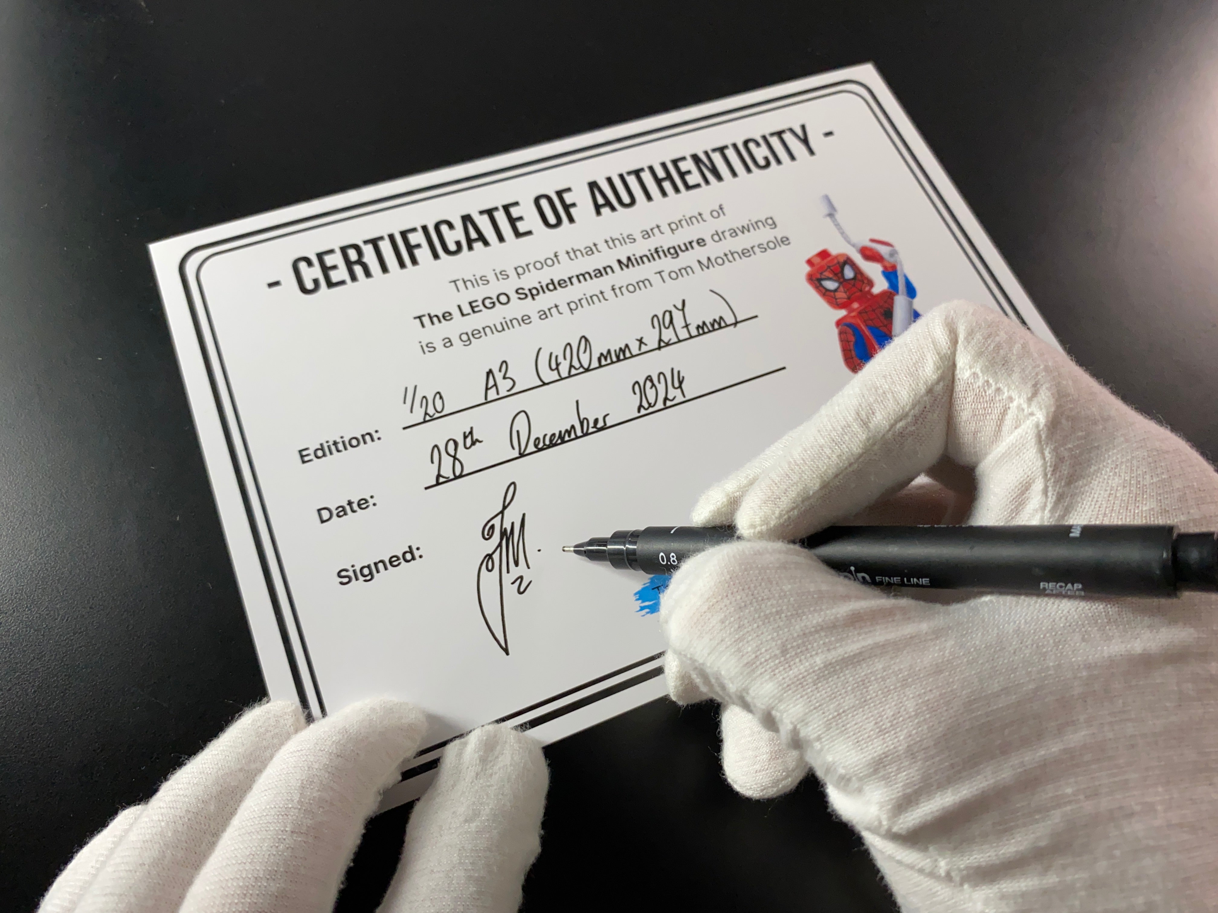 tom mothersole signing certificate of authenticity for realistic coloured pencil drawing of LEGO spiderman Minifigure